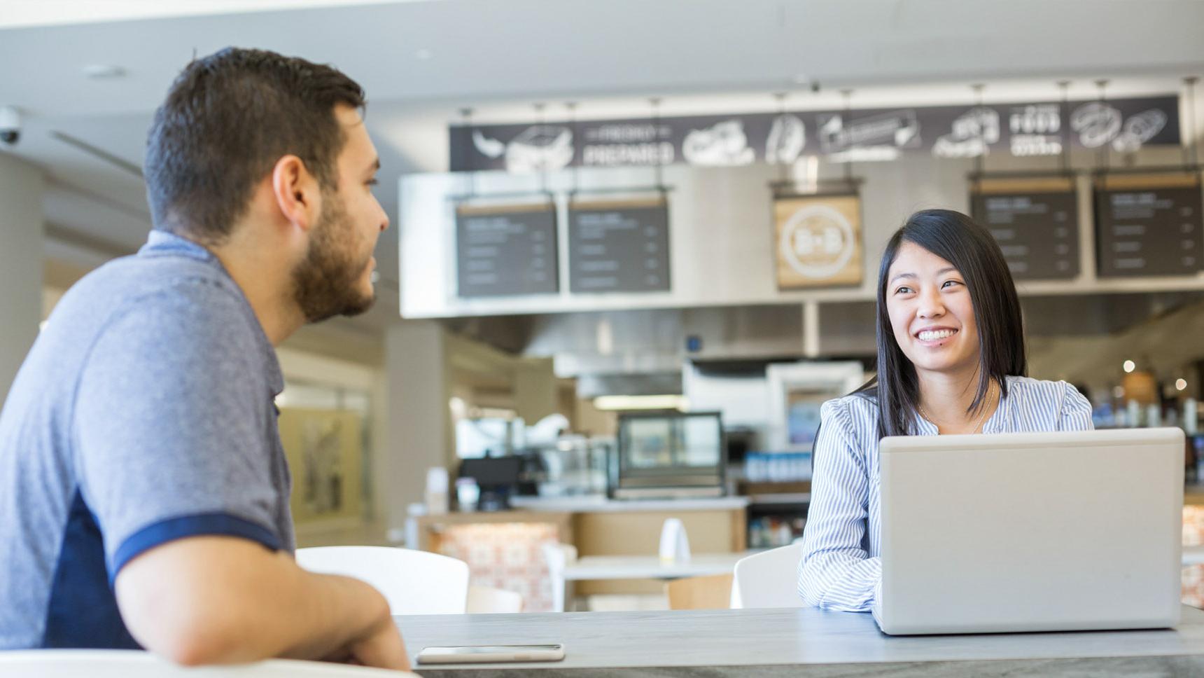 Two colleagues meeting in a cafe, working on their laptops, chatting and smiling