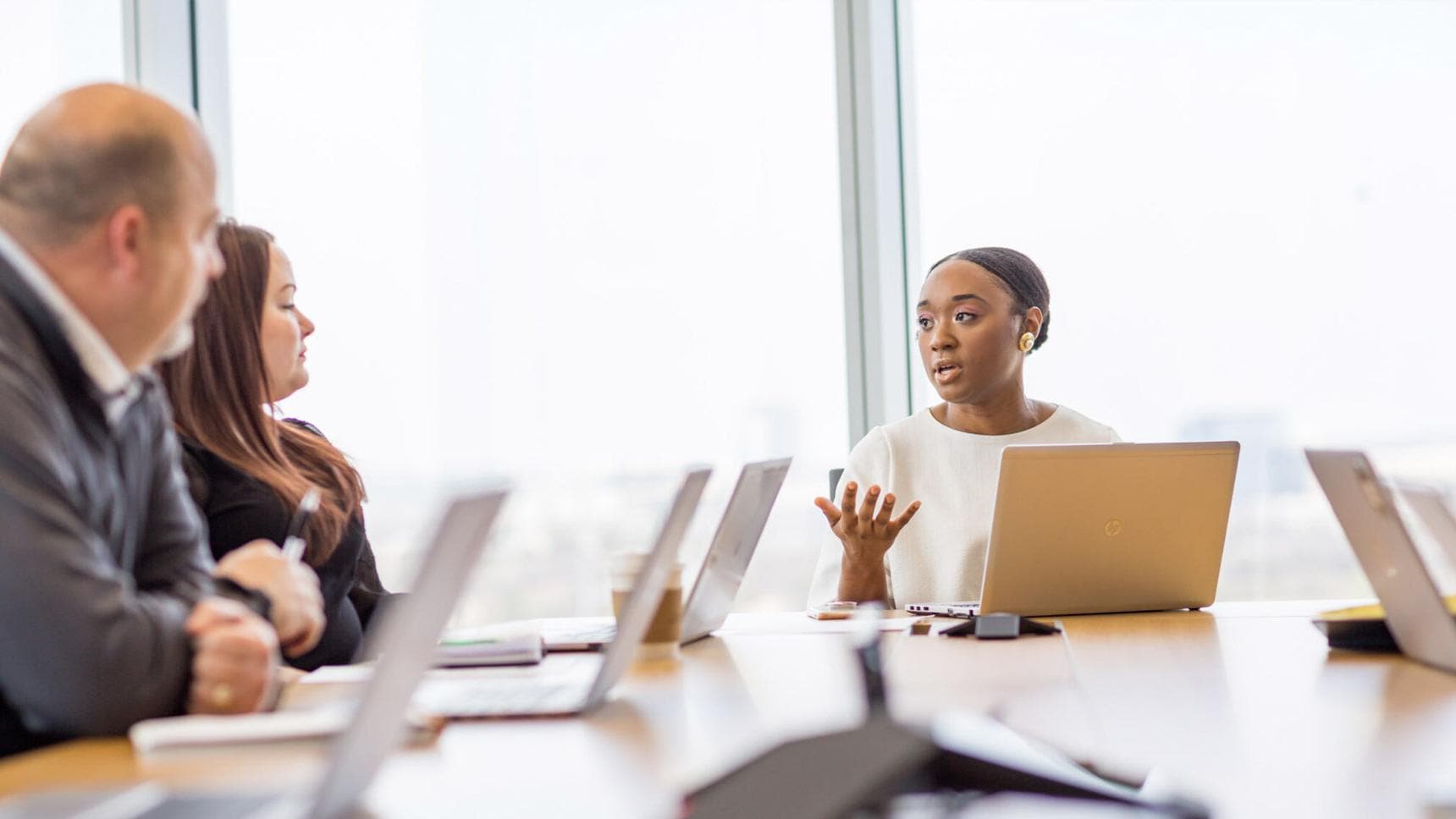 A diverse group of colleagues meeting in a conference room, all with laptops. One team member is at the head of the table seemingly presenting
