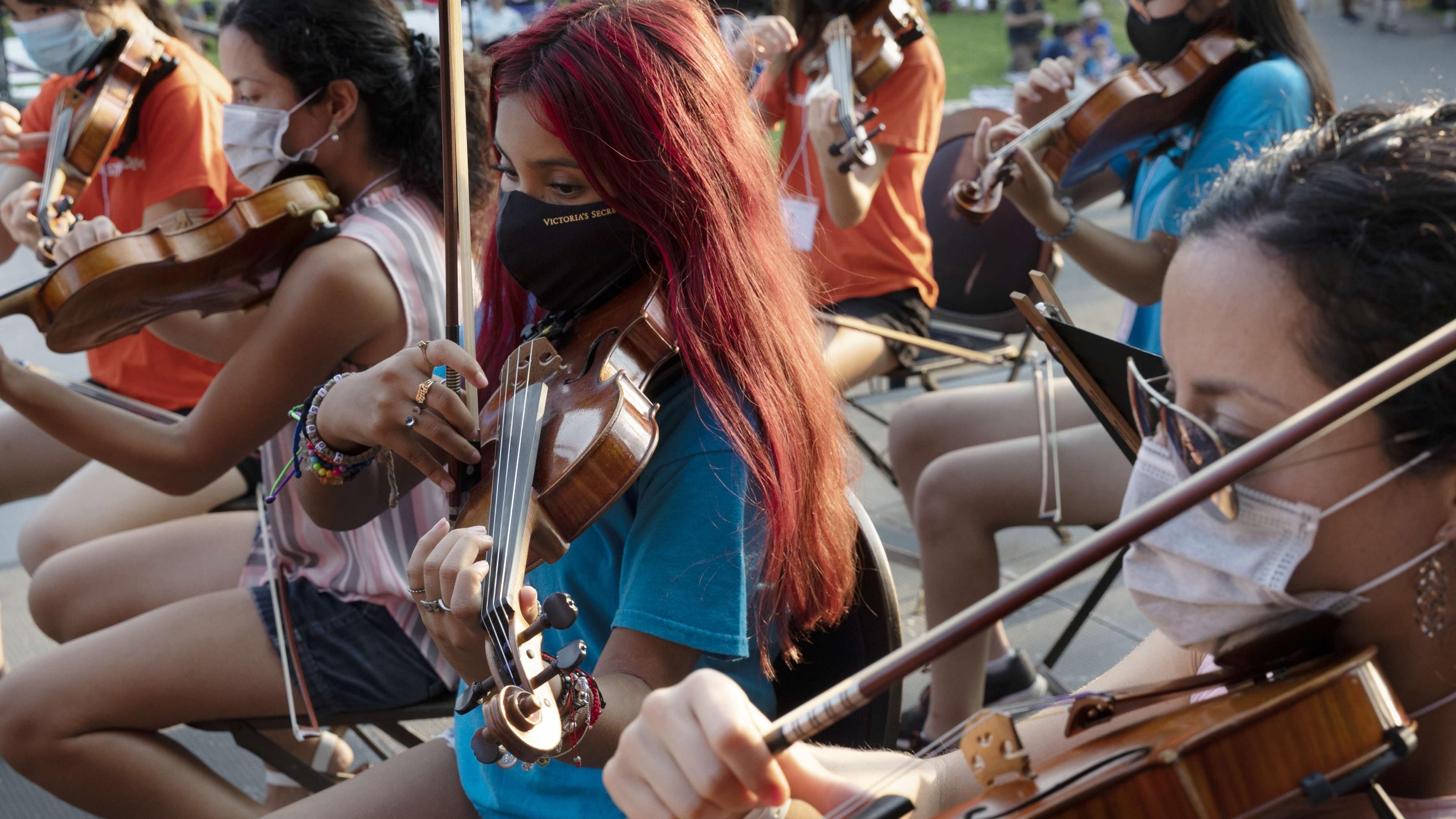 (slide 1 of 4) Masked young people playing the violin.