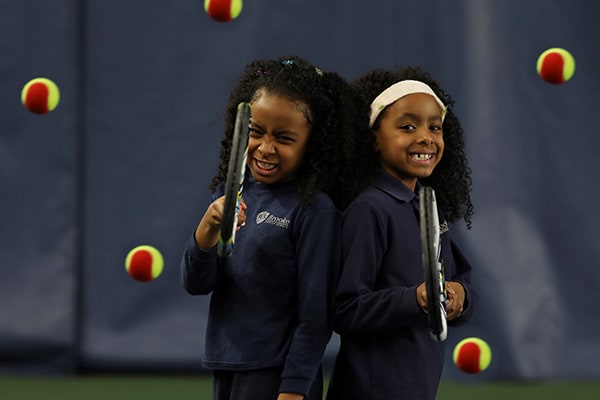 (slide 4 of 5) Two young children with tennis rackets standing back to back smiling. 